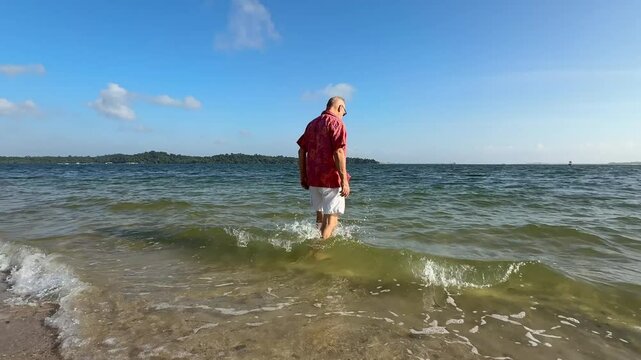 An elderly man, a senior citizen, strolls toward the deep ocean on a sunny day at a Singapore beach, enjoying the warm sand, gentle waves, and the vast blue sky above.