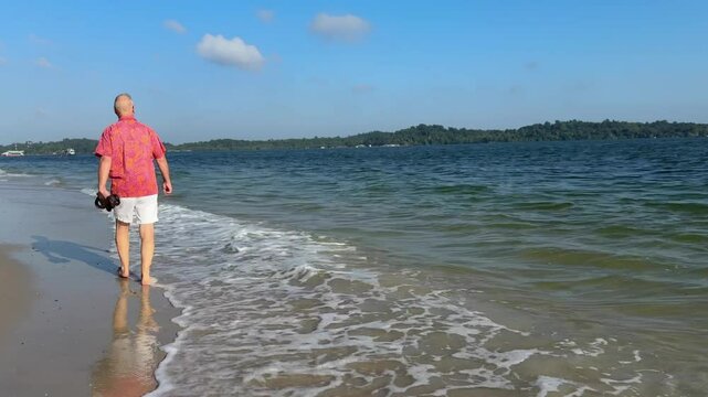 An man strolls along the seaside in Singapore, seemingly relaxing his feet as the cool ocean waves wash over them.