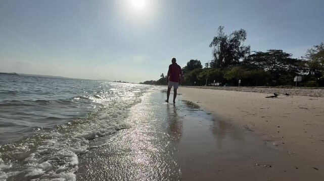 A silhouette of an elderly, senior citizen, European man walking on a sandy beach under a bright sunny day.