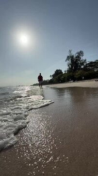 A peaceful beach scene captures gentle waves washing ashore under the bright sun. A solitary figure walks along the shoreline, with lush greenery and trees lining the background, evoking tranquility.