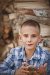 Portrait of a blond teenager. The boy is smiling. The teenager is wearing a checkered shirt. The boy is standing against the background of logs. A village boy. A boy on vacation. The boy is holding a 