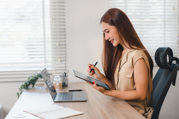 Woman working at desk with tablet and laptop, smiling in bright office environment.