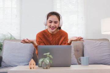 Woman wearing headphones sits on sofa, smiling and gesturing while video conferencing on laptop.