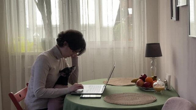 
Almaty, Kazakhstan - 02.02.2025 : A woman is sitting at her laptop, with her dog, in the kitchen. Work from home.