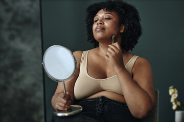 An African American woman using jade roller for facial massage while sitting beside makeup mirror...