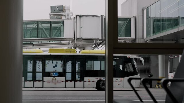 A public transportation bus is parked at an airport terminal, awaiting passengers. The environment features modern architecture and nearby boarding gates under overcast skies.