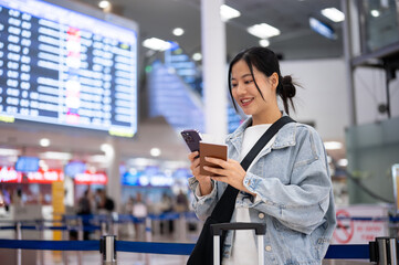 A happy Asian female passenger checks her flight boarding time on her phone, standing in the airport