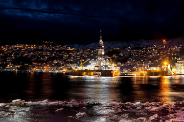 Fototapeta premium Brightly lit offshore rig against dark sky with illuminated town in background.