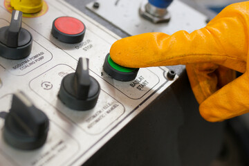 A worker wearing orange protective gloves pressing the green start button on an industrial control panel, initiating the operation of heavy machinery in a manufacturing facility.