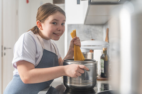 Beautiful, happy little teenage girl cooking in the kitchen at home. A girl in an apron puts spaghetti into a pan on a gray background.