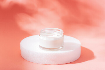 Glass jar of white cream on a white circular stand with a soft coral background, symbolizing skincare and beauty. Closeup