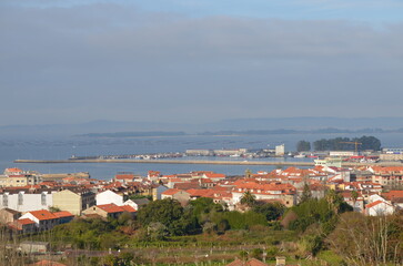 Coastal town with red-tiled rooftops overlooking a harbor, fishing boats, and floating mussel farms in calm blue waters, framed by distant hills and a soft morning sky
