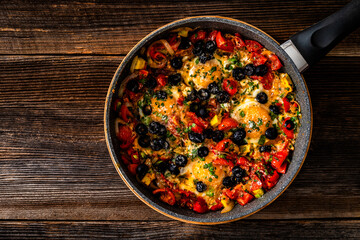 Shakshuka - fried eggs with vegetables and black olives in frying pan on wooden table