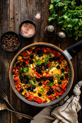 Shakshuka - fried eggs with vegetables and black olives in frying pan on wooden table