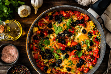 Shakshuka - fried eggs with vegetables and black olives in frying pan on wooden table