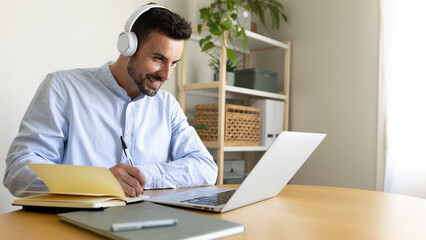 Fototapeta premium Happy man with headphones writing notes working at home using laptop. Copy space.
