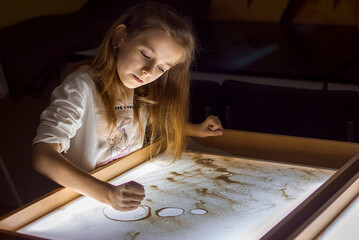 Beautiful little girl draws with sand on a light square, art therapy, sand animation, creative leisure for children, selective focus