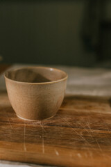 A bowl is sitting on a wooden cutting board. The bowl is brown and has a simple design. The cutting board is made of wood and has a few scratches on it. The scene is simple and uncluttered