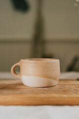 A white and tan coffee cup sits on a wooden cutting board. The cup is placed in the center of the board, and the background features a green wall. Concept of calm and relaxation