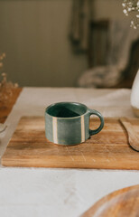 A green mug with a white stripe sits on a wooden cutting board. The mug is placed on a table with a spoon and a bowl nearby. Concept of warmth and comfort