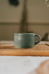 A green ceramic cup sits on a wooden table. The cup is small and has a handle. The table is made of wood and has a natural, rustic feel