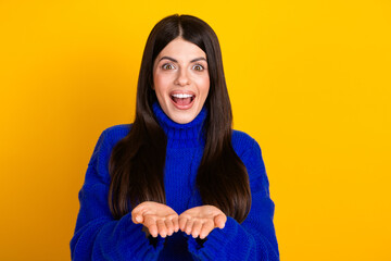 Joyful young woman presenting with open hands wearing cozy blue sweater in a bright yellow...