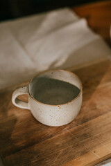 A white ceramic cup sits on a wooden table. The cup is empty and has a simple design. The table is made of wood and has a rustic feel to it. The scene is calm and peaceful