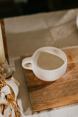 A white cup sits on a wooden cutting board. The cup is empty and the cutting board is made of wood