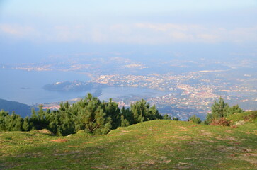 Panoramic view of Baiona's coastline from a lush, elevated viewpoint with green foreground, revealing a misty horizon and scattered clouds over the town and sea
