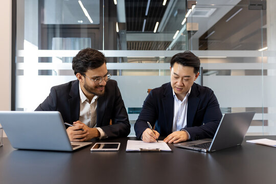 Two business people inside the office, signing a contract. Men in business suits inside the office, doing paperwork, sitting in a business meeting room.