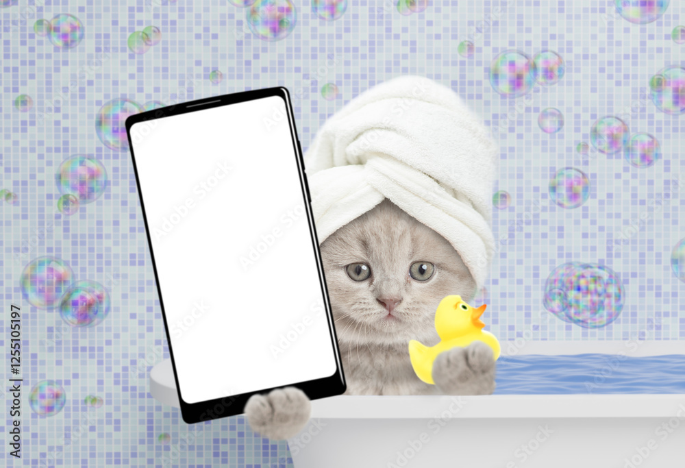 Poster Cute kitten with towel on it head takes the bath at home and shows big smartphone with white blank screen in it paw. Top down view