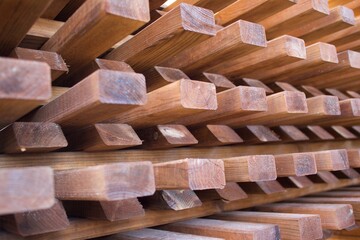 Close-up view of stacked wooden beams arranged in a geometric, grid-like pattern. The natural texture of the wood, with visible grain and knots, contrasts with the structured alignment, creating depth