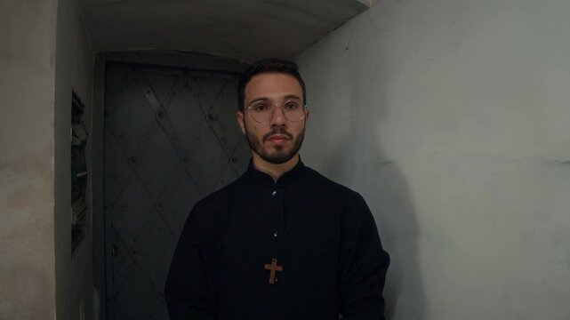 Waist-up portrait of confident young priest with facial hair wearing black cassock posing for camera holding bible book within church walls