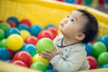 Asian baby A curious baby plays in a colorful ball pit, surrounded by bright plastic balls of various colors, looking up with wonder and joy.