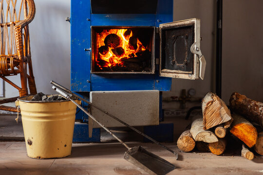 A blue solid fuel boiler with an open door reveals burning logs inside, emitting a warm glow. Stack of chopped wood is arranged on the floor. There are pieces of coal in a yellow metal bucket.