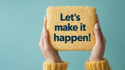 A person holds a textured yellow block with the motivational phrase "Let's make it happen!" against a light blue background.