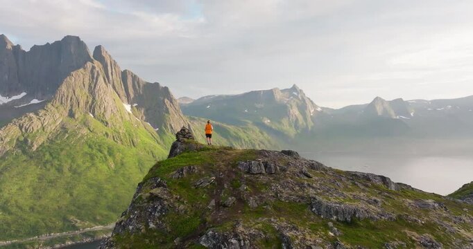 Hiker in arctic Senja overlooking jagged fjord mountain in sunset; drone reveal