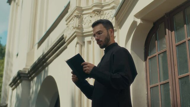 Low angle shot of concentrated young priest in black cassock reading holy bible outside catholic cathedral on sunny day
