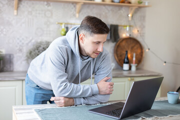 Smiling guy freelancer stand at kitchen desk read email message on laptop screen. Bearded...