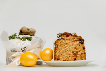 A festive Easter cake with white icing, decorated with green leaves, next to two vibrant yellow eggs and a wooden board, with a cut slice revealing a soft, fluffy texture with dried fruits inside.