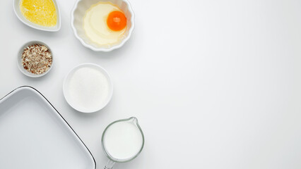 Flat lay composition for cooking preparation. Empty white baking dish with ingredients for baking, including egg, milk, butter, sugar, and nuts