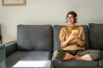 Happy smiling young student woman sitting on couch, listening music in headphones on mobile phone...