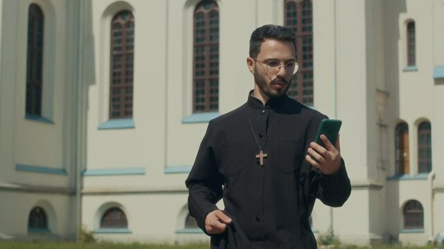 Young priest in black cassock with smartphone video calling relative and crossing them remotely while walking around church in summer