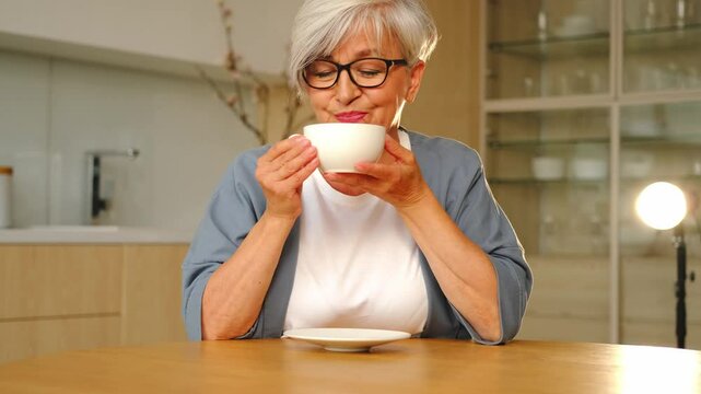 Good morning. Comfort relaxed senior old elderly woman grandmother drinking hot beverage tea coffee in kitchen at home. Mature woman resting enjoy calm morning at modern home alone. Pension concept