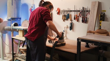 Craftsperson works with orbital sander with sandpaper on lumber to achieve smooth finish, checking quality. Woodworking expert in carpentry shop using angle grinder to fix wooden surface, camera B