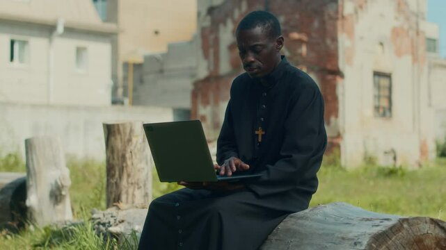Modern African American priest dressed in black cassock working on laptop while sitting on log on summer day