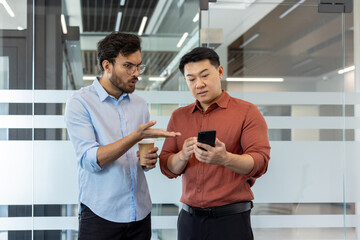 Two colleagues debating and analyzing data on a smartphone while standing in a professional workspace