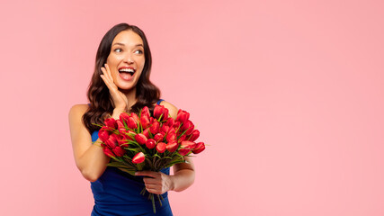 Overjoyed young lady with red tulips bouquet feeling excited, looking aside at free space on pink studio background. Woman's Day offer, spring holiday concept