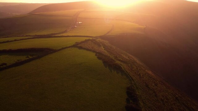 Descending Sunset Aerial View Over Coastal Fields in Devon with Bright Setting Sun Flare and Hazy Background. Drone Rural Farming Footage 4K.