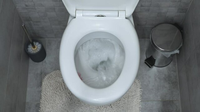 A top-down view of a toilet being flushed in a modern bathroom. The scene includes a toilet brush, a small trash bin, and a textured bath mat, creating a clean and organized space.